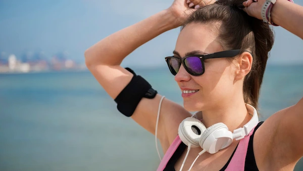 Mujer deportista al aire libre usando lentes de sol negros y audífonos frente al mar.